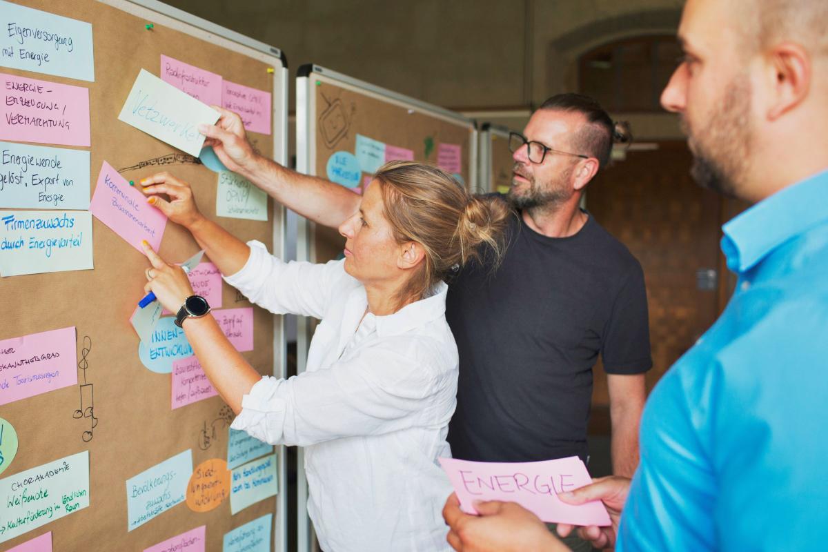 Drei Personen pinnen gemeinsam beschriebene Zettel an eine bereits gut gefüllte Pinnwand