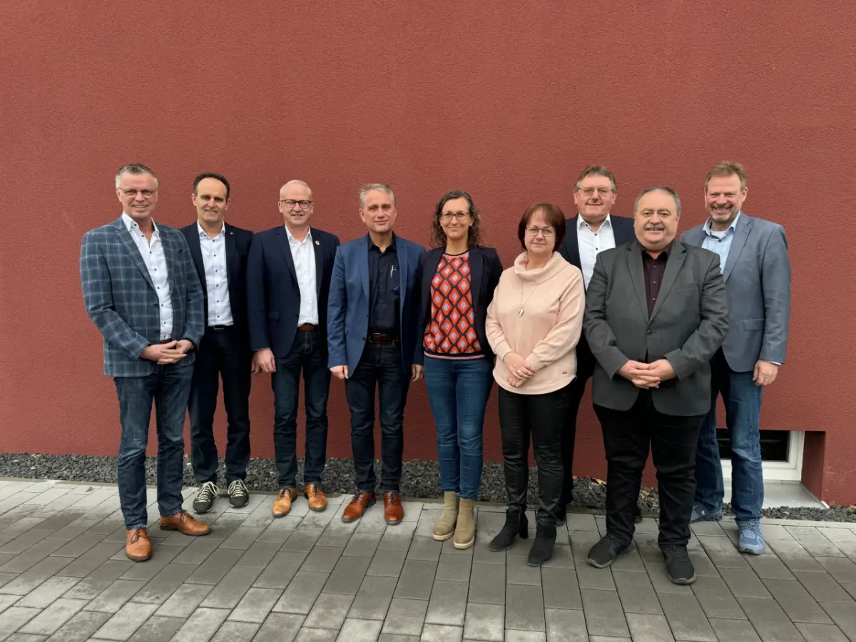 Sieben Männer und zwei Frauen stehen vor roter Wand und sehen in die Kamera.