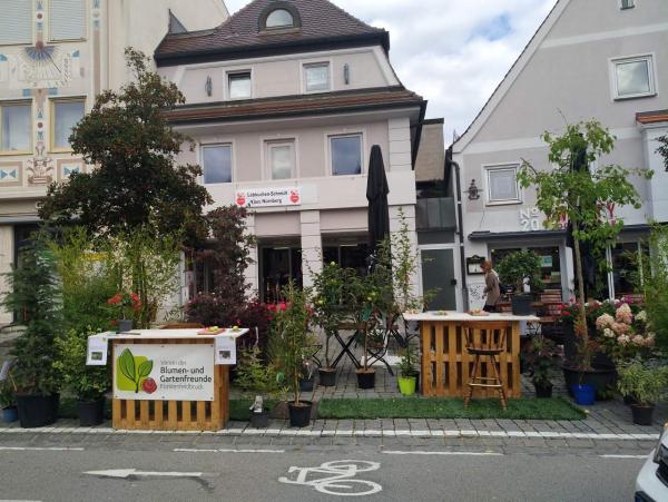 Im Vordergrund eine Fahrradstraße. Dahinter eine umgenutzte Parkplatzfläche mit Sitzgelegenheiten. Dahinter drei Häuser in einer Reihe.