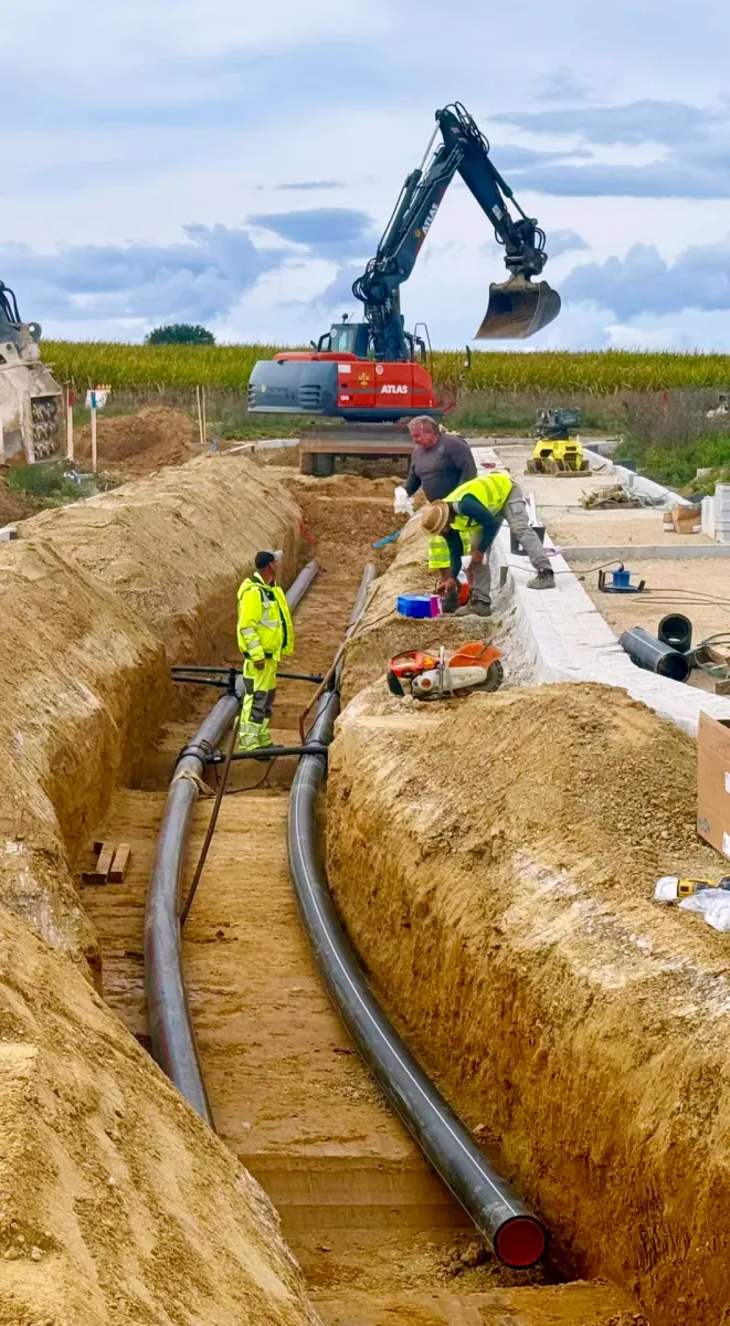 Drei Menschen verlegen zwei schwarze Rohre in einem Graben.