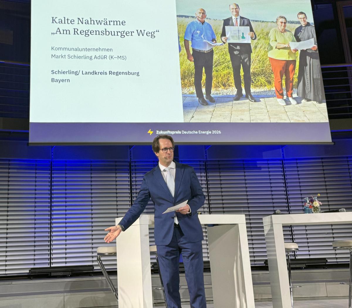 Mann steht auf Bühne. Im Hintergrund PowerPoint Präsentation.