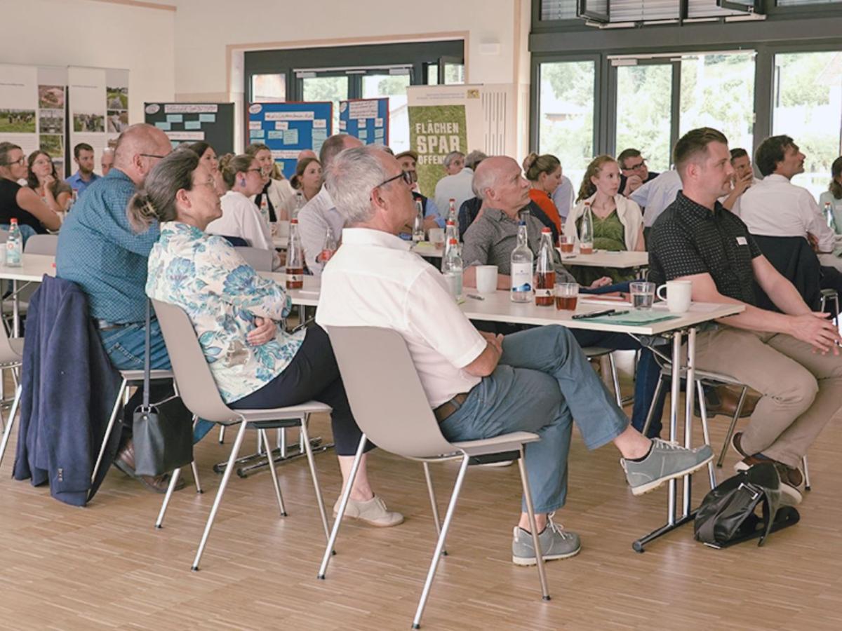 Personen sitzen an Tischen und sehen nach rechts zum Vortrag