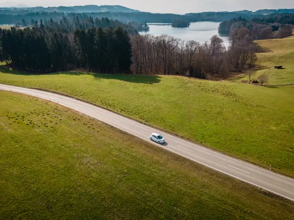 Weißes Auto fährt auf einer landstraße. Im Hintergrund ein Wald und ein See bei schönem Wetter.