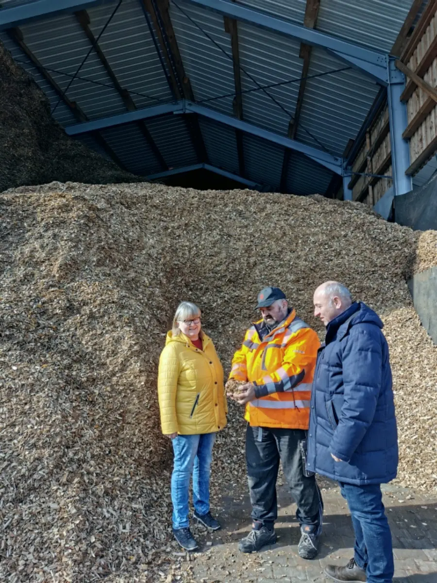 Zwei Männer und eine Frau stehen vor einem Berg mit Hackschnitzeln und sehen sich den Brennstoff an.