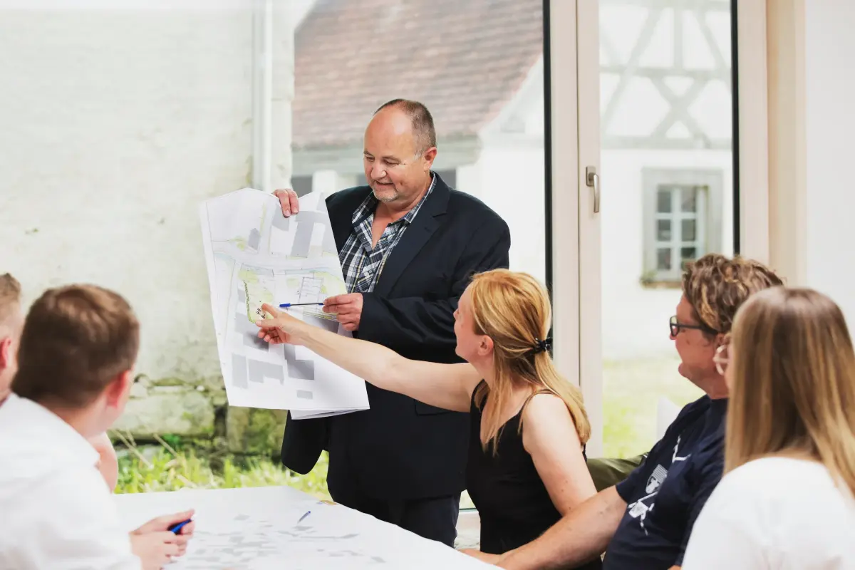 Mehrere Personen sitzen am Tisch zusammen. Ein Mann hält einen Plan hoch, er und eine Frau zeigen darauf. Durch das Fenster im Hintergrund ist ein Fachwerkhaus zu erkennen.