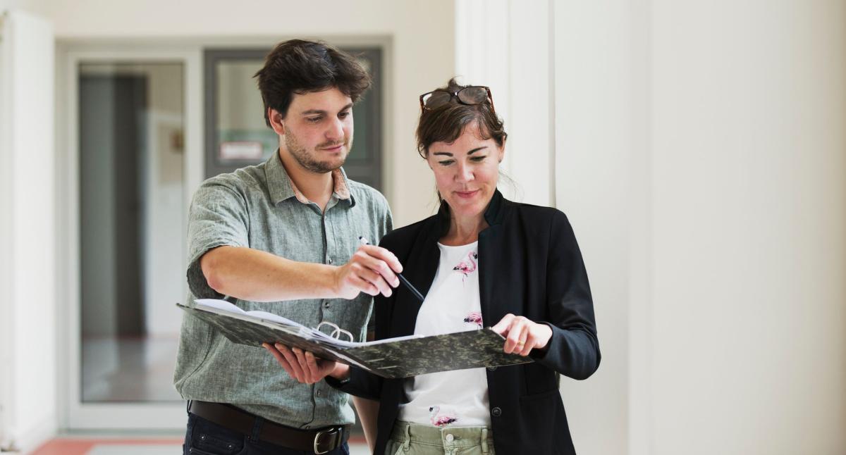 Eine Frau und ein Mann stehen in einem langgestreckten, hellen Flur mit hohen Decken zusammen. Die Frau hält einen aufgeschlagenen Aktenordner, in den sie gemeinsam blicken.