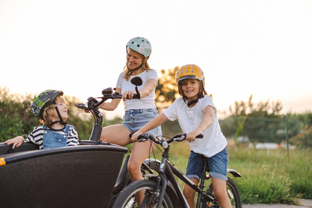 Mutter mit zwei Kindern. Das größere Kind fährt ein Fahrrad. Mutter fährt mit kleinerem Kind Lastenfahrrad. Szene im ländlichen Raum.