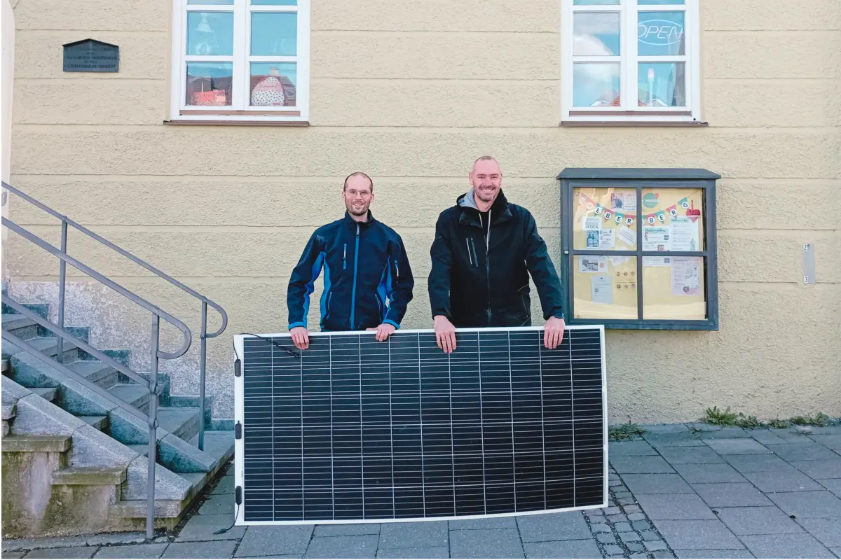 Zwei Männer stehen vor einer gelben Hausfassade, halten ein Solarpanel und lachen in die Kamera.