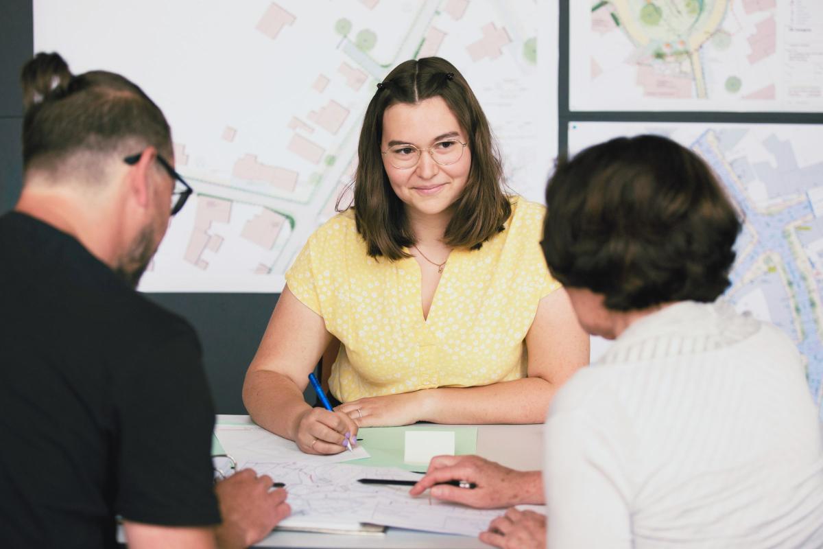 3 Personen sitzen an einem Besprechungstisch mit Plänen und Unterlagen zusammen, im Hintergrund sind Pläne angepinnt